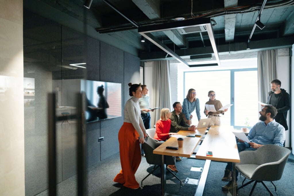 A group of eight people gather around a conference table in a modern office, networking and discussing documents and laptops, as natural light streams in from a large window in the background.