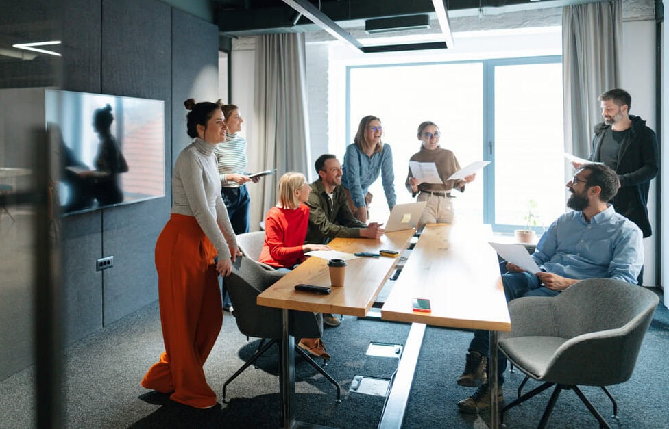A group of eight people gather around a conference table in a modern office, networking and discussing documents and laptops, as natural light streams in from a large window in the background.