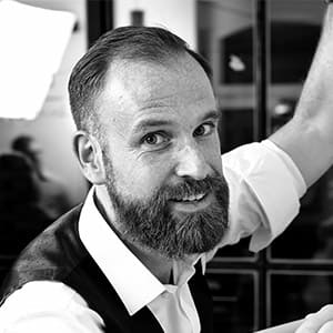 A bearded man with short hair, wearing a white shirt and a dark vest, smiles at the camera in a black and white photo. He raises one arm, and a blurred background suggests an indoor setting.