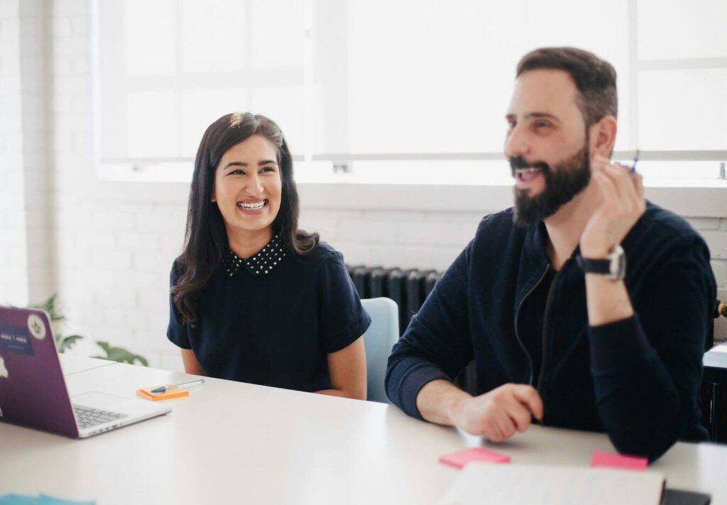Two people sit at a white desk in a bright office, smiling and talking about networking tips. A laptop, papers, and pens are on the table. The atmosphere appears friendly and collaborative.