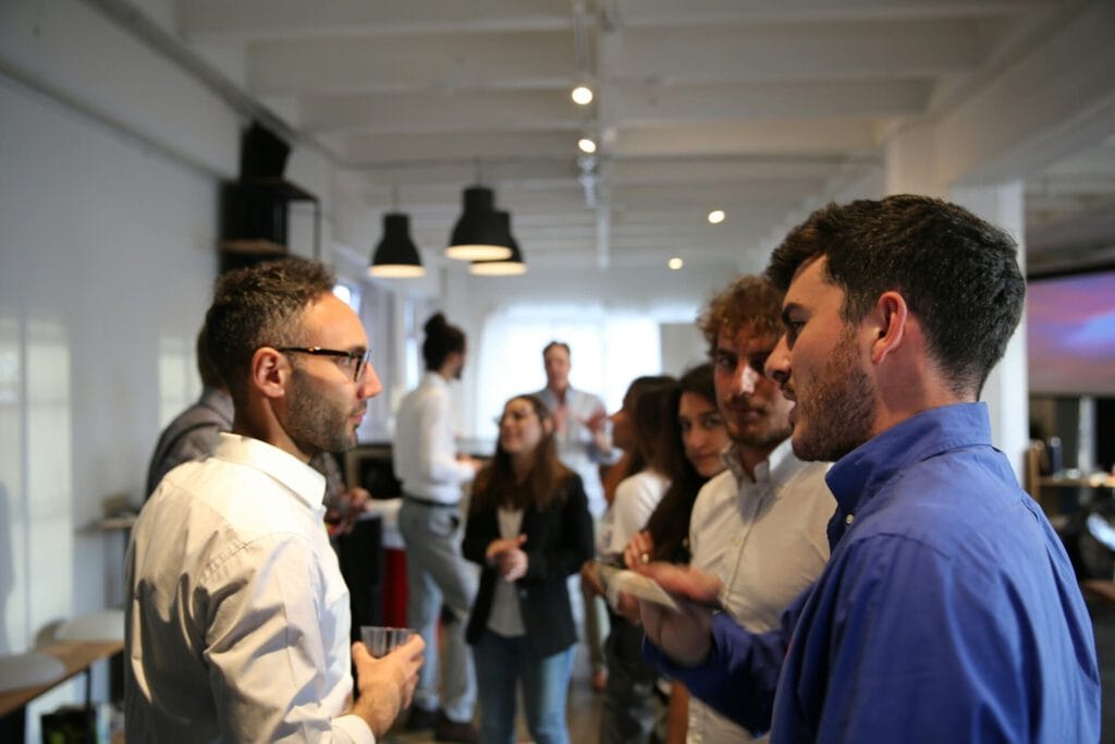 A group of people are gathered indoors, engaged in conversation. Several individuals in the foreground are talking and holding drinks, while others interact in the background. The setting appears to be a casual social or networking event.