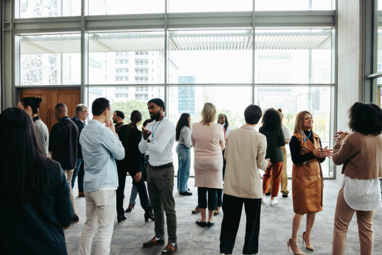A group of people stand and mingle in a bright, modern room with large windows, engaging in conversation and networking in small groups.