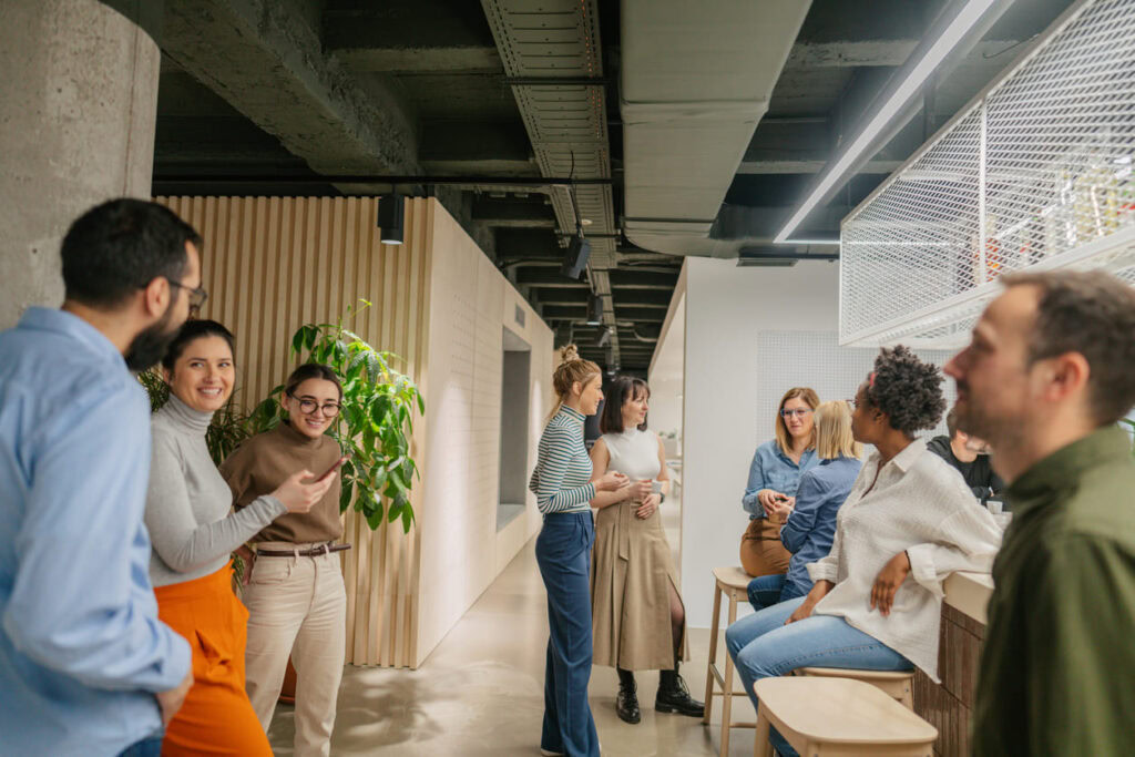 A group of people casually chatting and smiling in a modern office space with light wood accents and plants. Some are standing, others are sitting on stools near a counter.