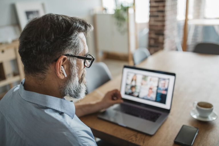 A man with glasses and earbuds sits at a table using a laptop for a video call. A smartphone and a cup of coffee are on the wooden table, and the room is softly lit with natural light from a window.