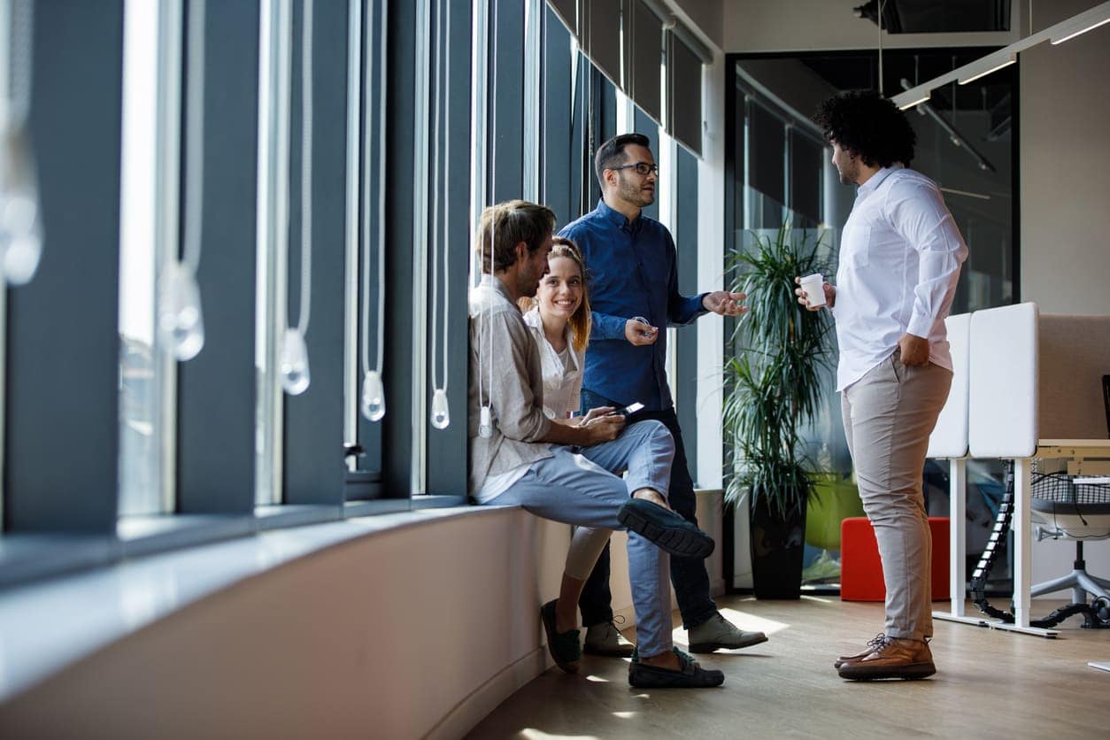 Four people casually chat about personal development in a bright office corridor by large windows. Two sit on a ledge, one stands with a phone, and another holds a coffee cup. They appear relaxed and engaged in conversation.