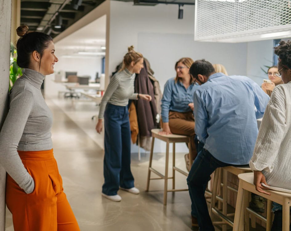 A group of people in casual office attire are gathered around a high table, talking and laughing, while one woman stands against a wall, smiling and observing the group. The setting is a modern, open-plan office.