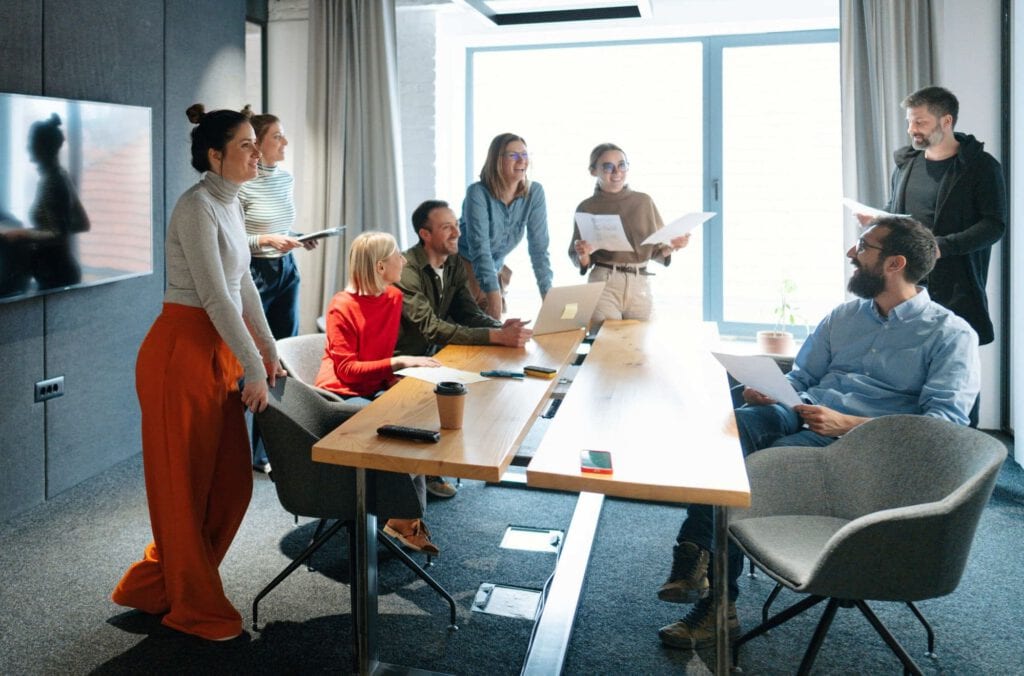 A group of eight people, five standing and three seated, have a discussion around a conference table during corporate events, with papers, a laptop, and coffee cups in a modern, well-lit office meeting room.