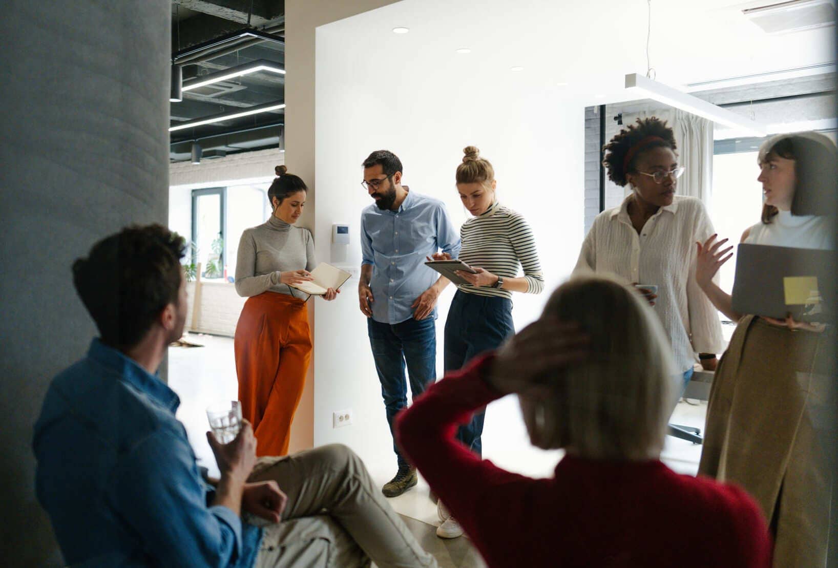A group of six people stand and talk in a modern office space, some holding notebooks or tablets, while two more sit nearby engaged in a discussion about the 2025 European Automotive landscape and its potential industry impact.