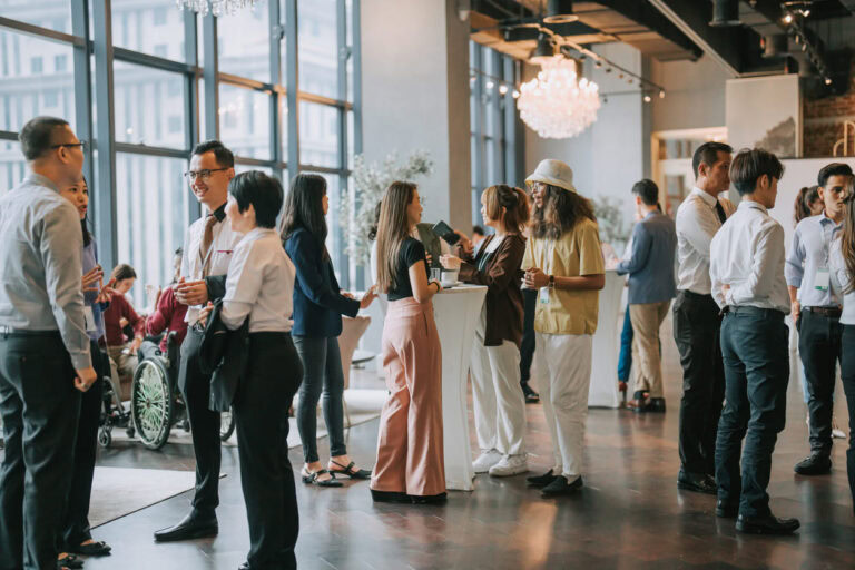 A group of people in business attire mingle and converse at a networking event in a modern, well-lit indoor space with large windows and a chandelier. Some hold drinks and there is a person in a wheelchair present.